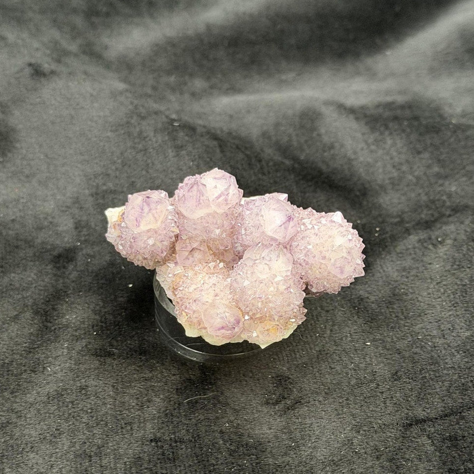 A cluster of pale pinkish-lavender crystals on a small clear display stand, placed on a dark charcoal velvet surface.